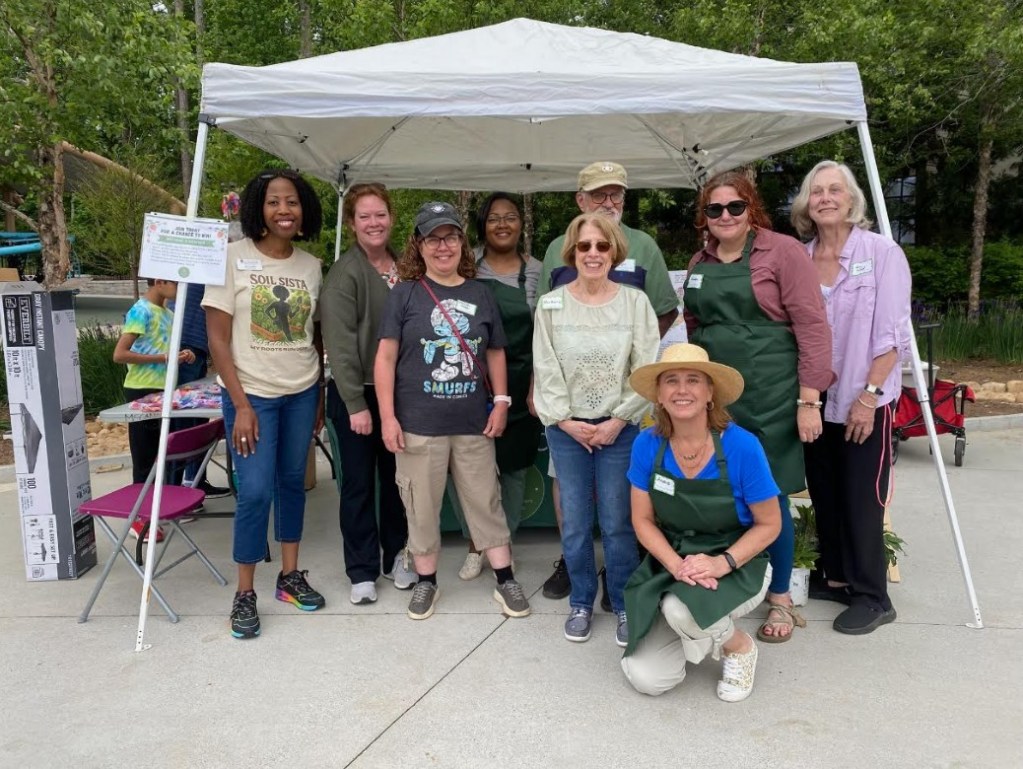 A group of nine women and one man posing together under a canopy tent, featuring greenery in the background. They are wearing casual clothing, with some in green aprons, and smiling at the camera. Tables with supplies are visible nearby, suggesting a community or gardening event.
